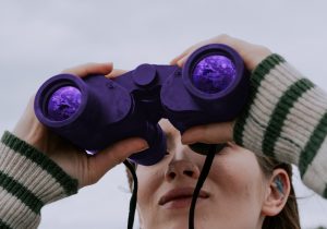 Person looking through binoculars with purple-tinted lenses representing vision, planning, and strategic foresight for business development and future planning.