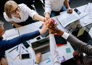 Business team putting hands together in teamwork gesture over desk with laptops, charts and documents representing collaboration and team building in professional workplace.