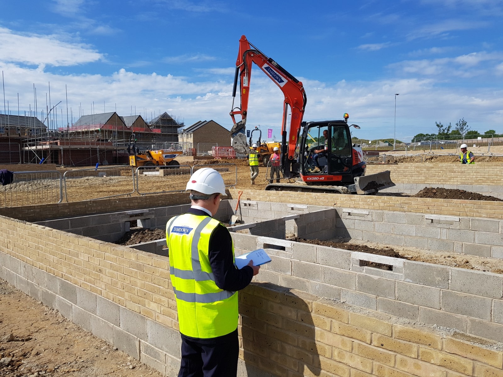 Construction site supervisor in hi-vis jacket and hard hat conducting quality inspection of concrete block foundations with excavator and residential housing development in background.